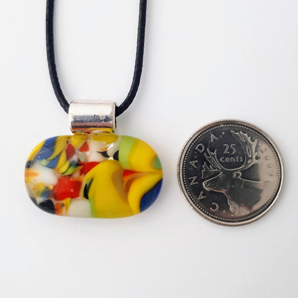 Wide oval glass fused necklace pendant with colourful blue, red, yellow, with small bits of green and white chunks/specs in it, with silver coloured bail and black cord on white background with a quarter for scale