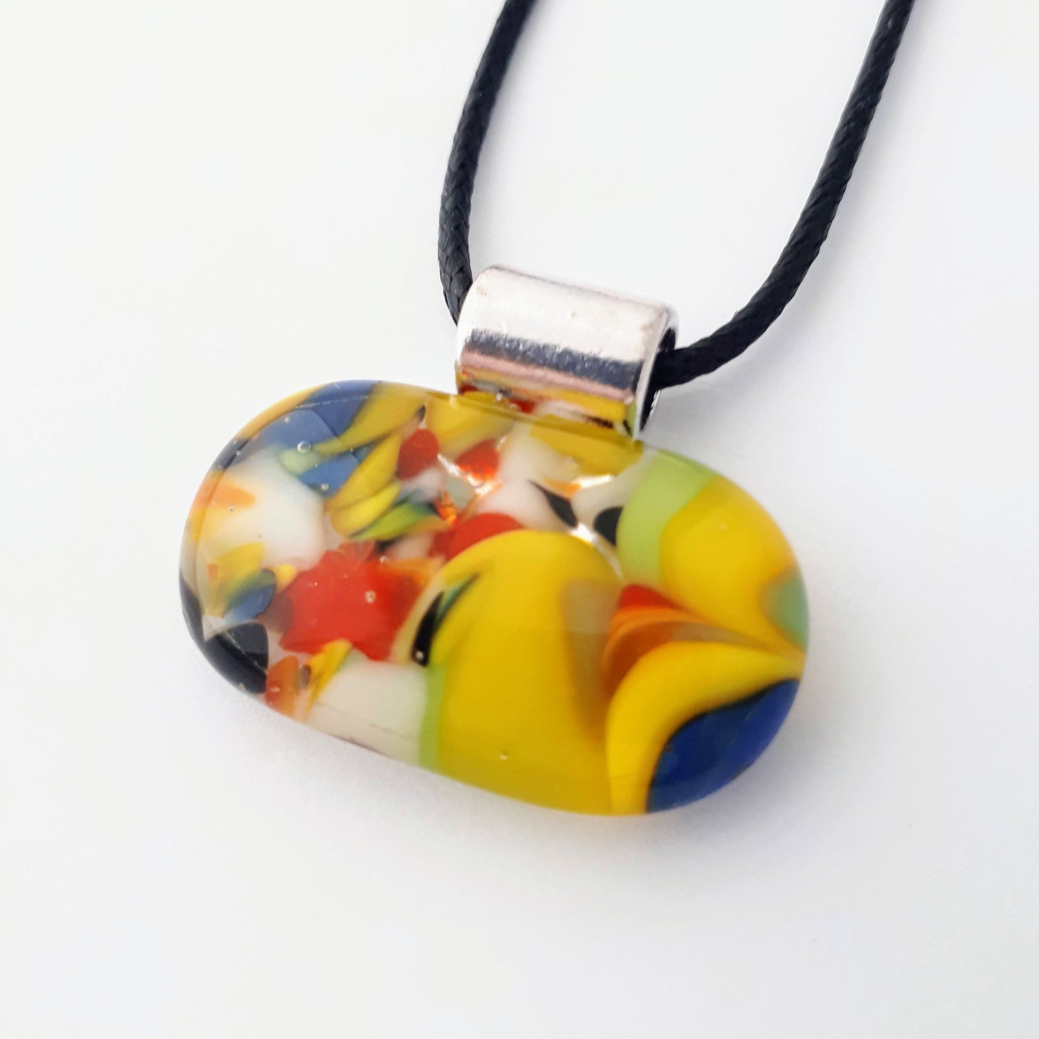 Wide oval glass fused necklace pendant with colourful blue, red, yellow, with small bits of green and white chunks/specs in it, with silver coloured bail and black cord on white background
