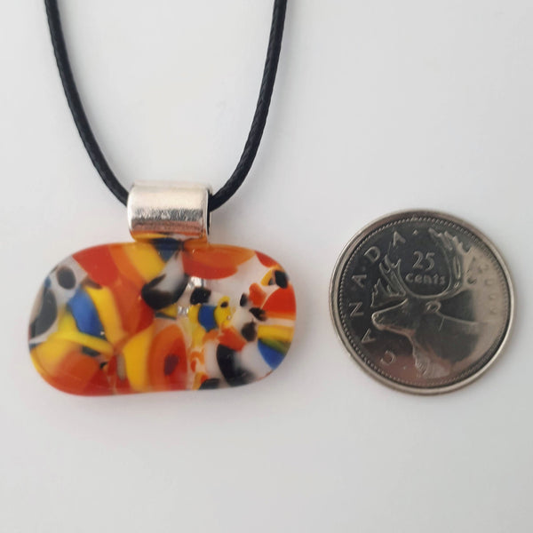 Wide oval glass fused necklace pendant with colourful blue, red, yellow, with small bits of green and white chunks/specs in it, with silver coloured bail and black cord on white background with a quarter for scale