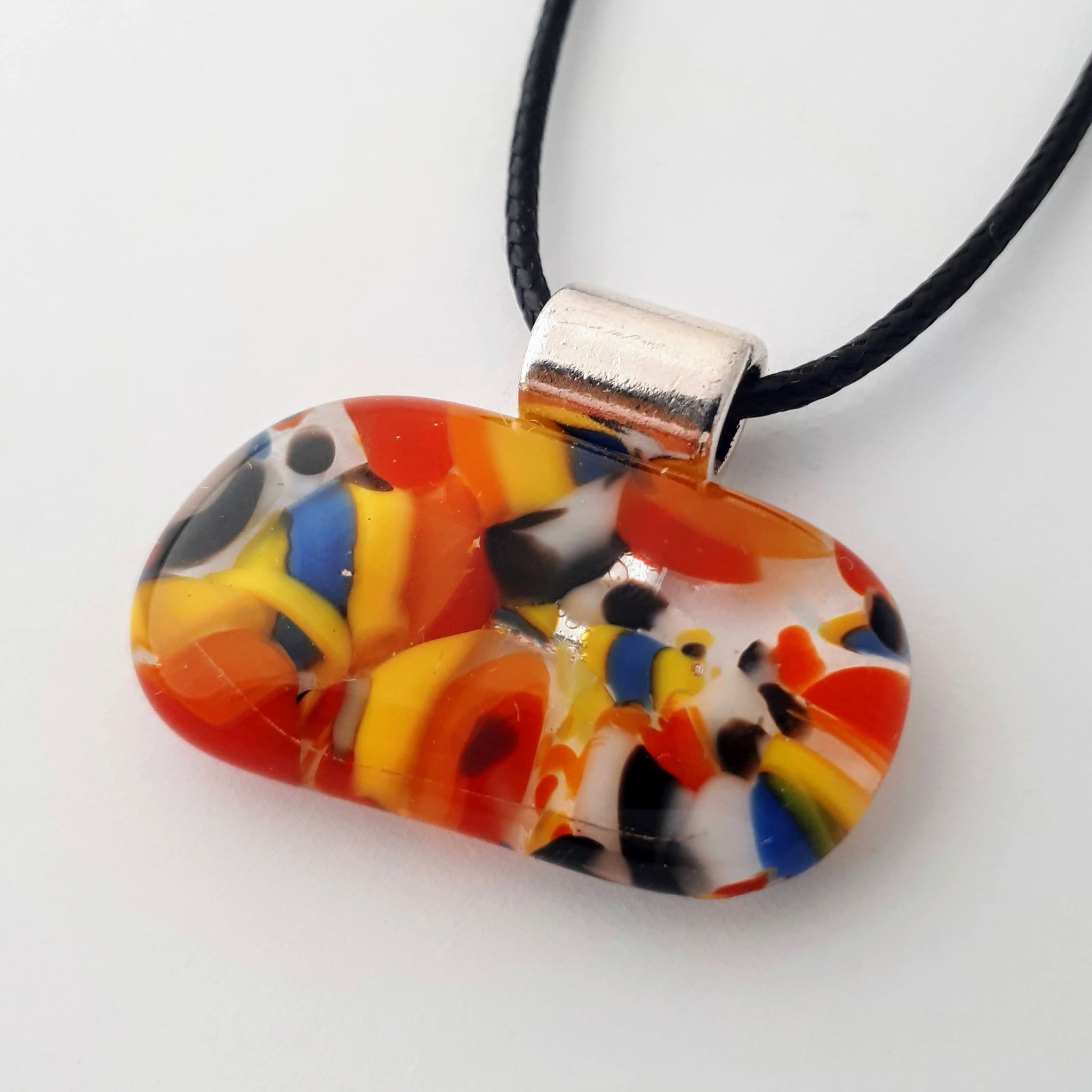 Wide oval glass fused necklace pendant with colourful blue, red, yellow, with small bits of green and white chunks/specs in it, with silver coloured bail and black cord on white background