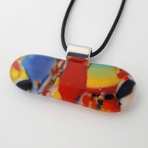 Wide oval glass fused necklace pendant with colourful red, blue, yellow, with small bits of green, black and white chunks/specs in it, with silver coloured bail and black cord on white background