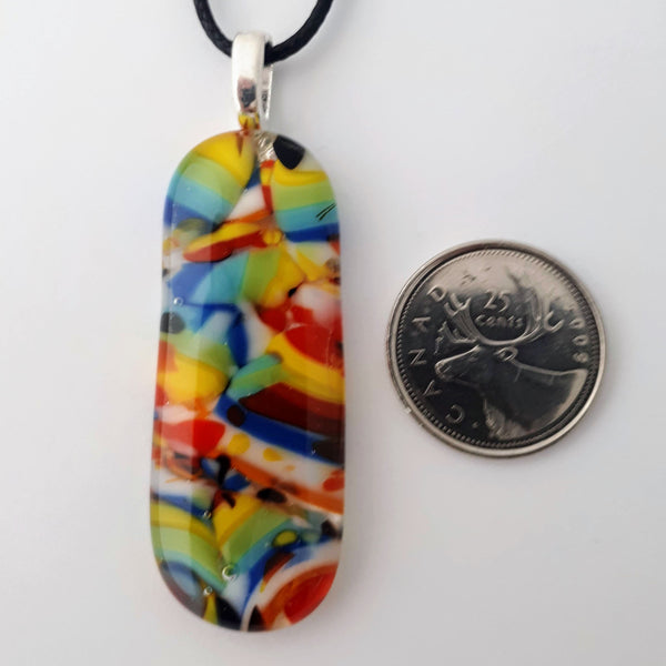 Long oval glass fused necklace pendant with colourful blue, red, yellow, with small bits of green and white chunks/specs in it, with silver coloured bail and black cord on white background side view with quarter for scale