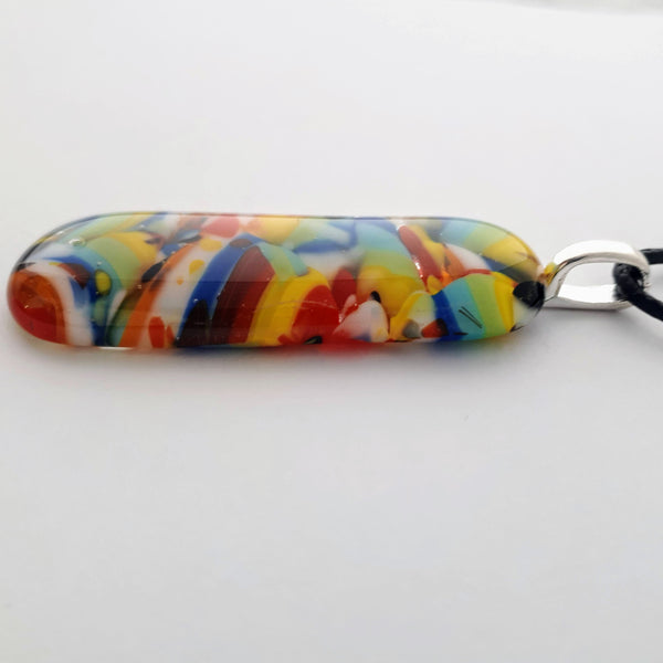 Long oval glass fused necklace pendant with colourful blue, red, yellow, with small bits of green and white chunks/specs in it, with silver coloured bail and black cord on white background side view