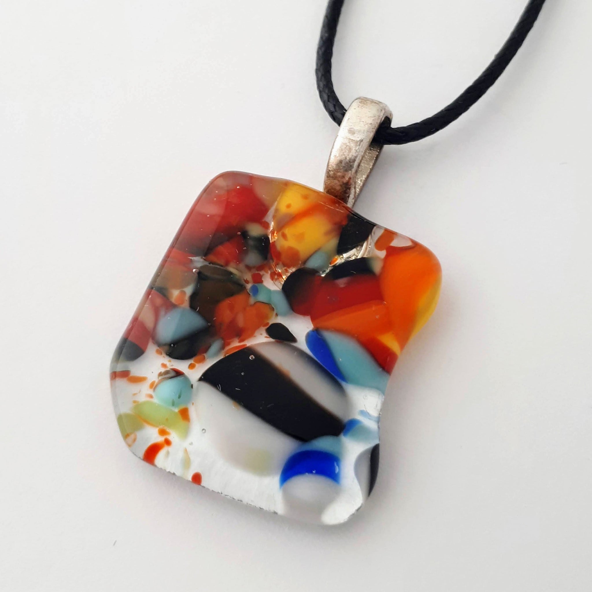 Square-ish glass fused necklace pendant with colourful red, orange, black, white, and bits of blue/green chunks/specs in it, with silver coloured bail and black cord on white background