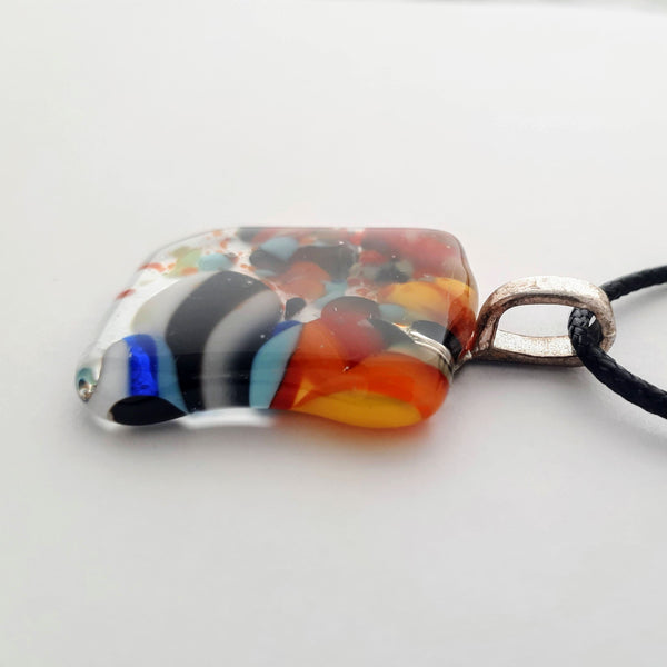 Square-ish glass fused necklace pendant with colourful red, orange, black, white, and bits of blue/green chunks/specs in it, with silver coloured bail and black cord on white background side view