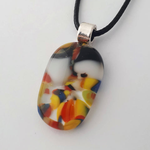 Vertical oval glass fused necklace pendant with colourful black, white, red, yellow, with small bits of blue and green chunks/specs in it, with silver coloured bail and black cord on white background