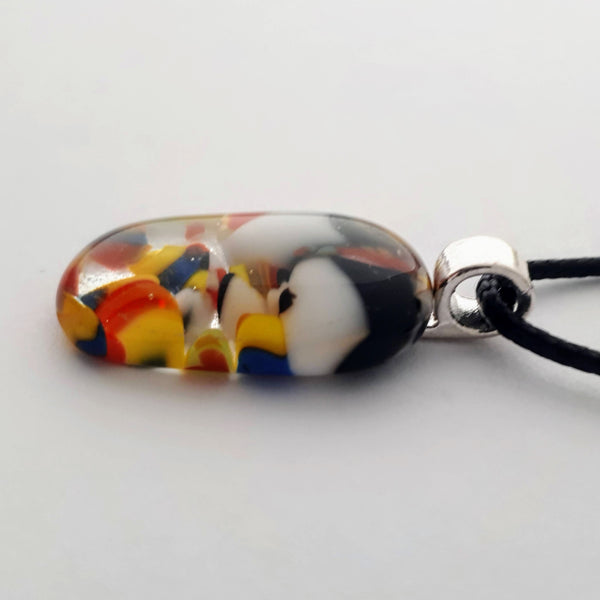 Vertical oval glass fused necklace pendant with colourful black, white, red, yellow, with small bits of blue and green chunks/specs in it, with silver coloured bail and black cord on white background side view