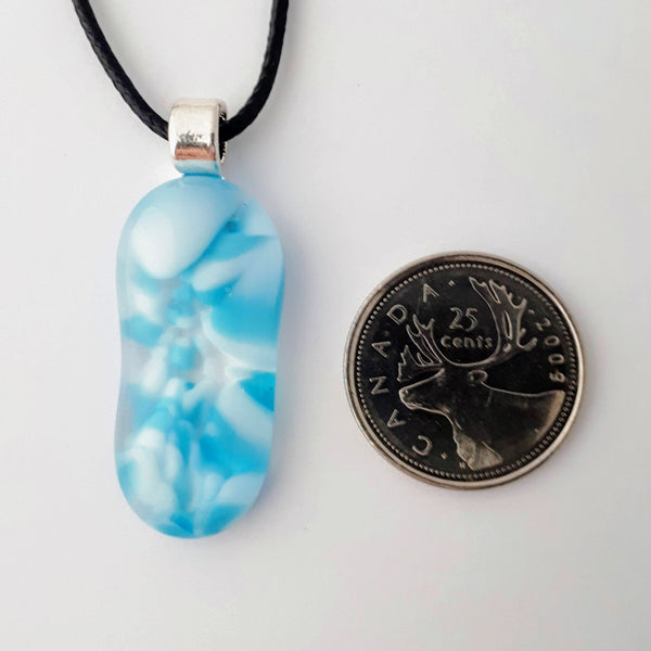 Blue and white glass fused pendant, long oval, with silver bail and black cord on white background with quarter for scale
