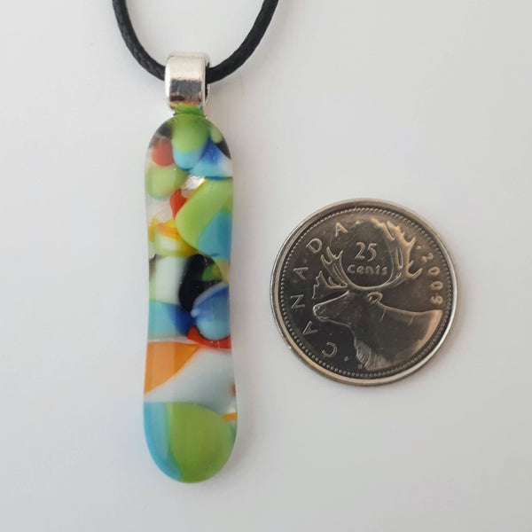 Mainly blue and green multicoloured long skinny glass necklace pendant on white background with black cord and silver coloured bail with quarter for scale