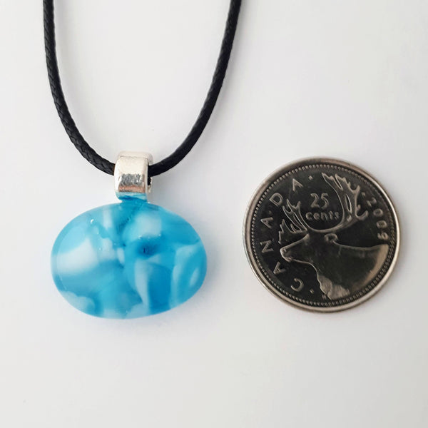 Blue and white wide oval glass fused pendant with silver bail and black cord on white background with quarter for scale